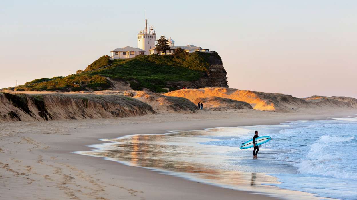 Surfer walking into the surf at Nobbys Beach with their surfboard, close to Nobbys Lighthouse