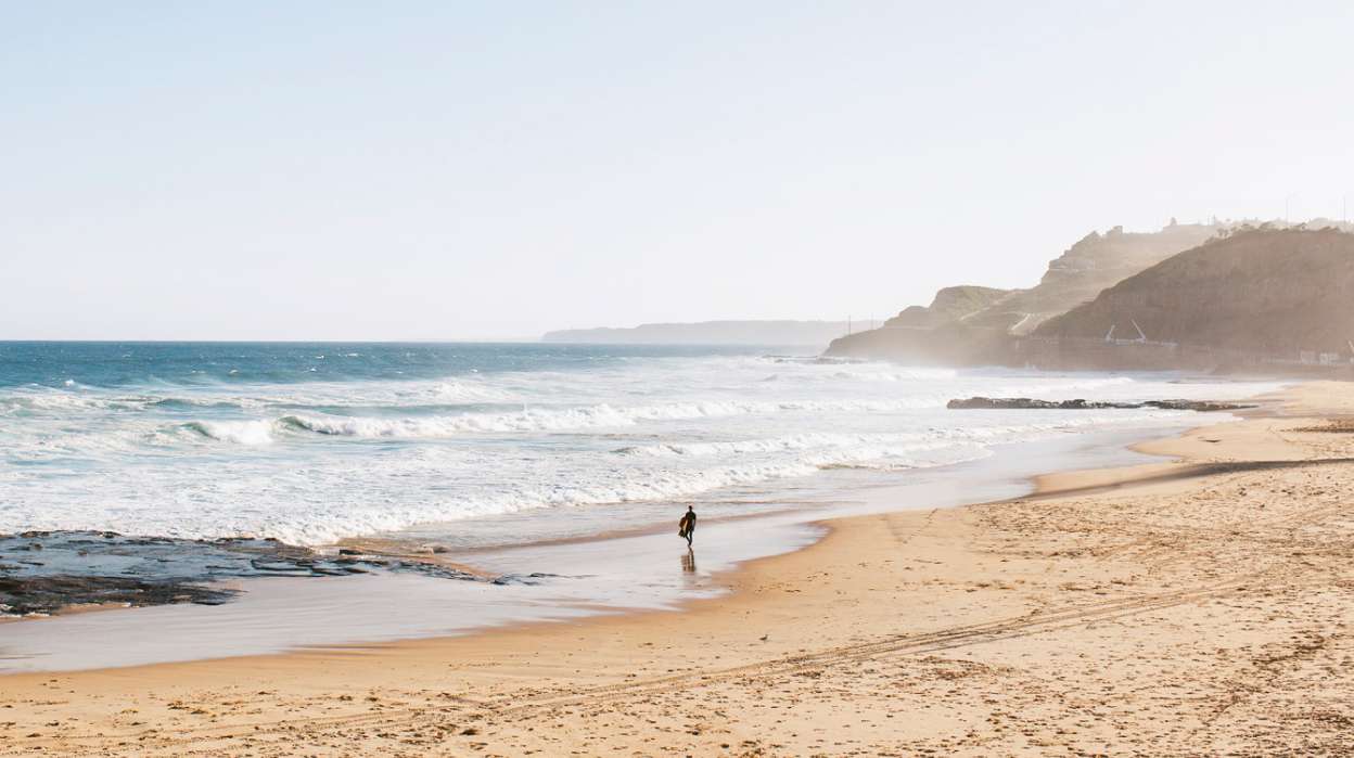A surfer walking on an empty Newcastle Beach, with the coastline in the distance